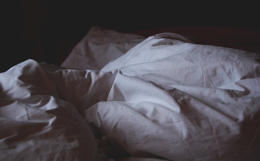 Picture of white bedding & pillows crumpled up on top of a bed like someone just got out from under the covers. Background of photo is all black.