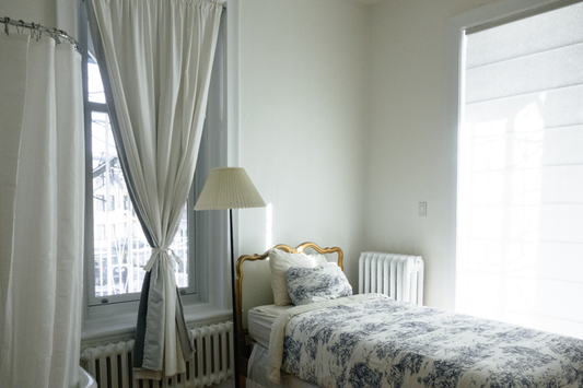 Twin bed in corner of a room with a lamp next to it and two windows. One covered by sheer window blinds and the other covered by a white curtain tied in the middle and draping down.