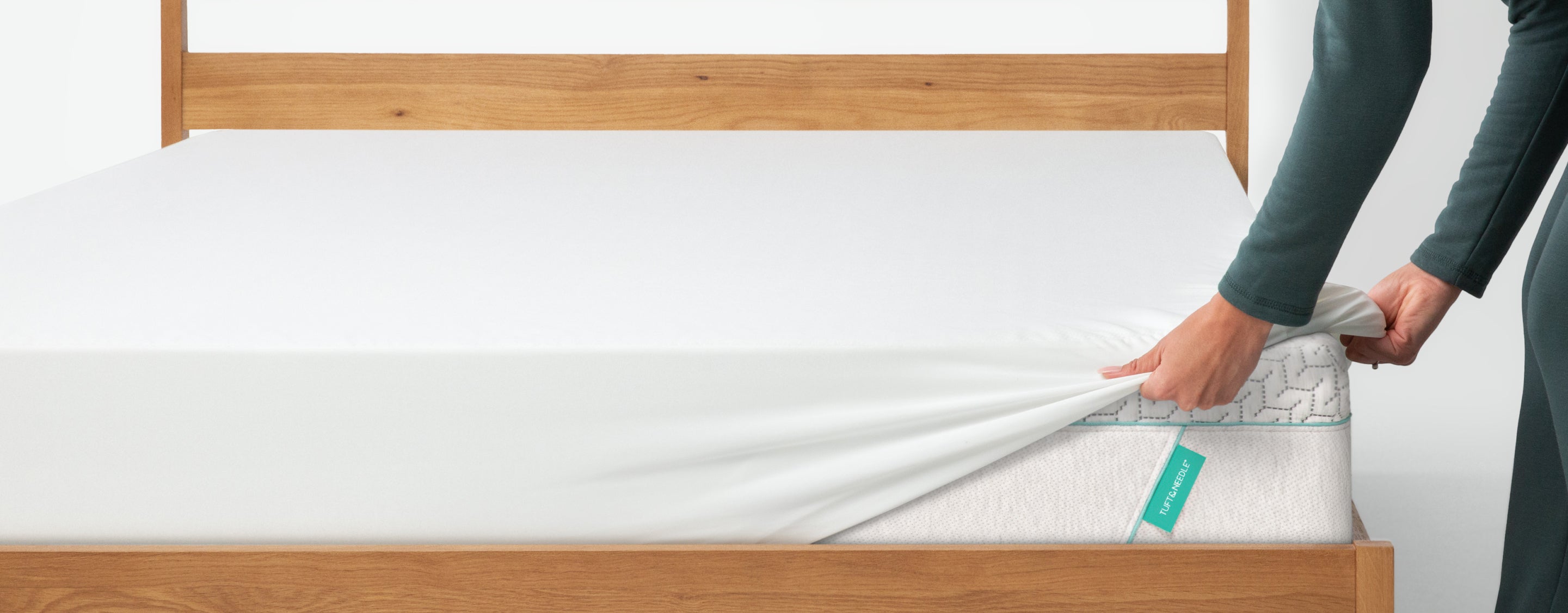 A person's hands and arms are shown pulling a white fitted sheet off the corner of a Tuft and Needle mattress on a wooden bed frame. The mattress has a small green tag attached to it.