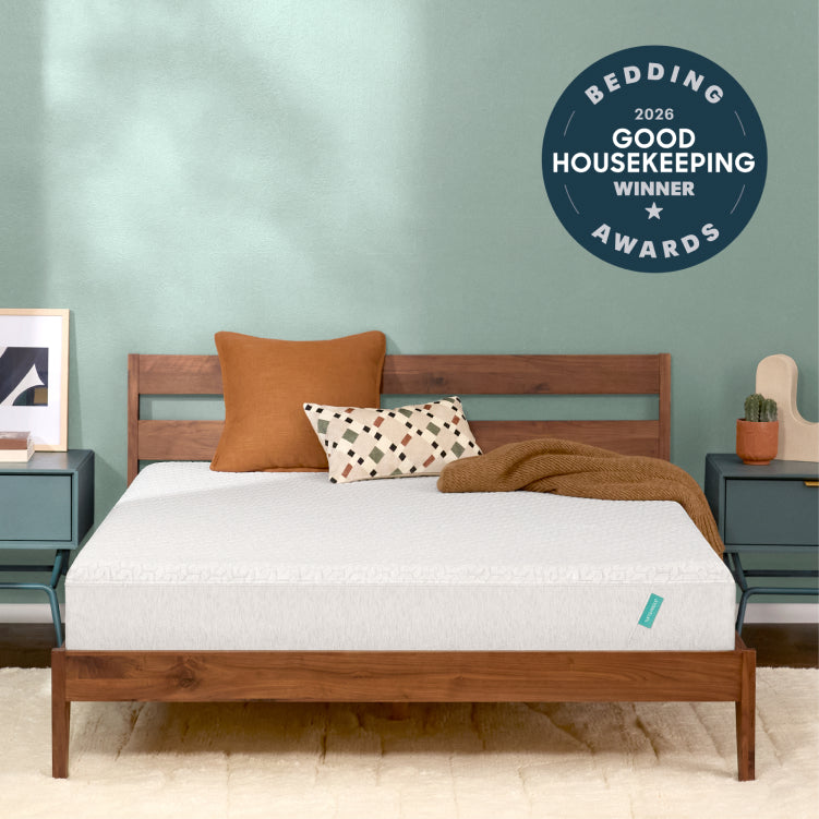 A Tuft and Needle Original mattress, styled with two pillows and a brown throw blanket, set against a sage green wall. A dark blue circular badge in the top right reads 'BEDDING 2026 GOOD HOUSEKEEPING AWARDS WINNER'.