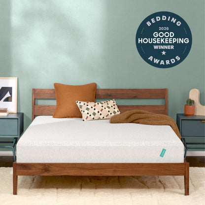 A Tuft and Needle Original mattress, styled with two pillows and a brown throw blanket, set against a sage green wall. A dark blue circular badge in the top right reads 'BEDDING 2026 GOOD HOUSEKEEPING AWARDS WINNER'.