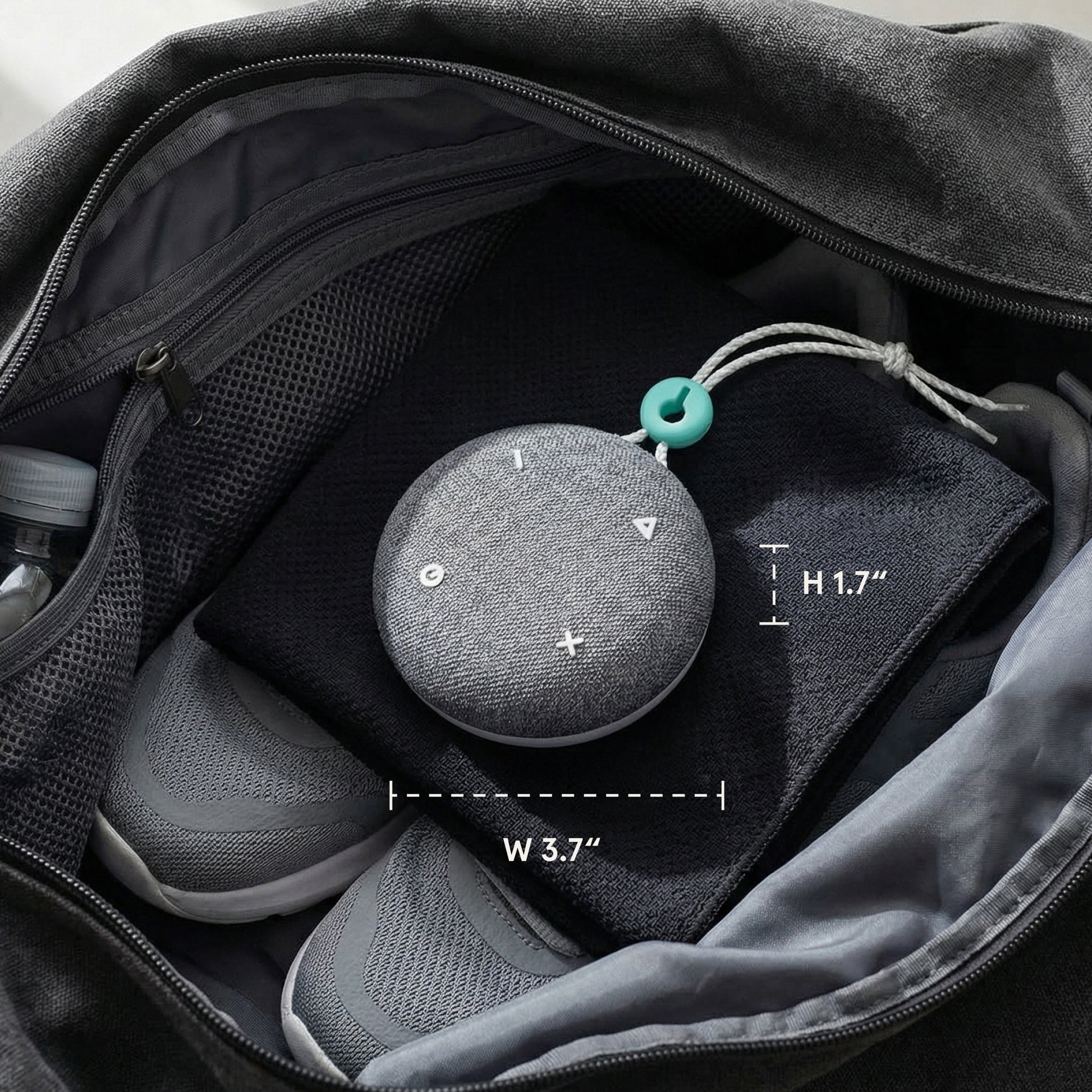 A Tuft and Needle SNOOZ Go 2 Travel White Noise Sound Machine, round gray fabric speaker, with white control symbols and a small teal hanging ring, shown inside an open black bag with size annotations reading 3.7 inches wide, 1.