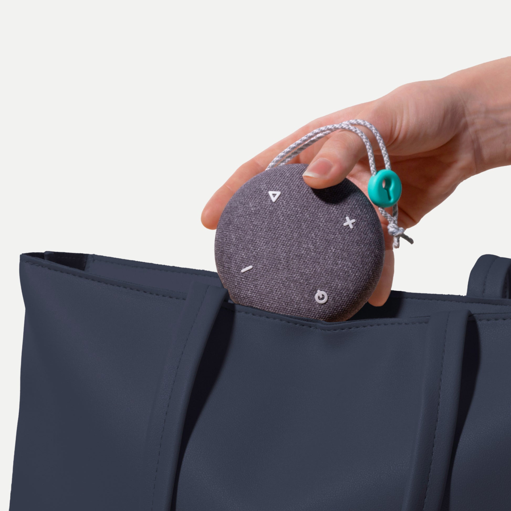 A hand holding a small Tuft and Needle SNOOZ Go 2 Travel White Noise Sound Machine, round gray, with a silver cord and turquoise bead, shown near a navy tote bag against a light neutral background.