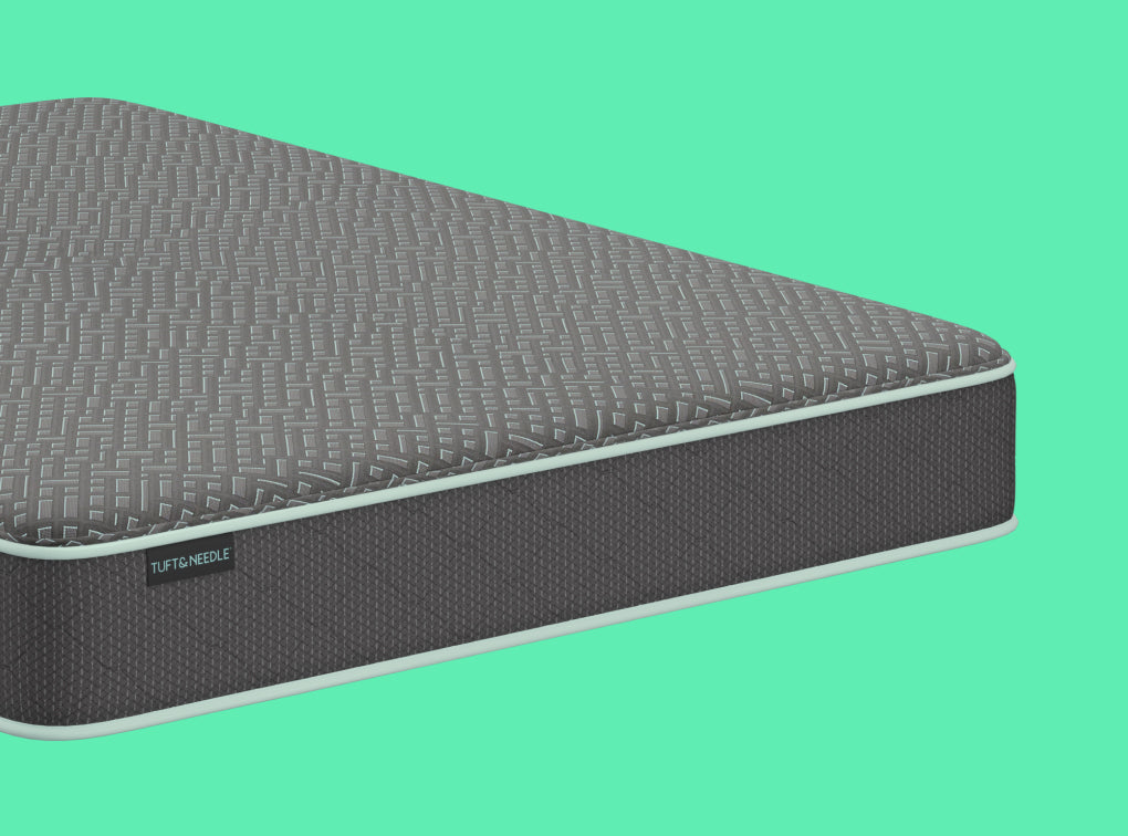A close-up of a Tuft and Needle Essential mattress corner showing a textured gray top surface and a dark charcoal side panel with a small visible TUFT & needle brand tag, set against a solid mint green background.