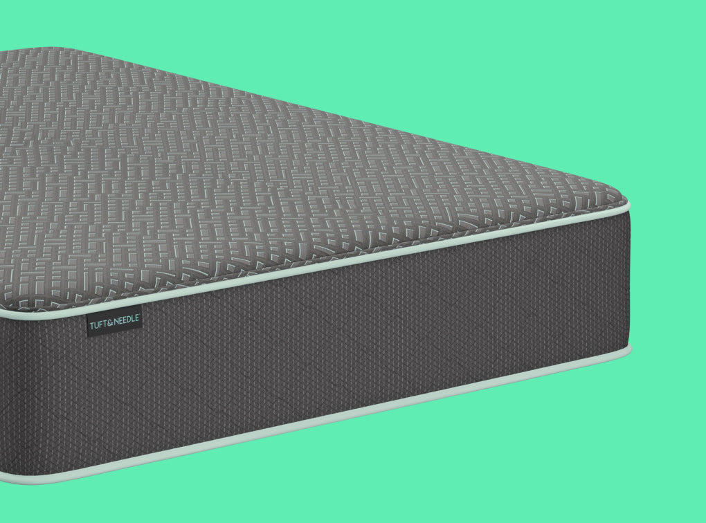 A close-up of a Tuft and Needle Mint II mattress corner showing a textured gray top surface and dark gray side panel with a small visible TUFT & needle brand tag, set against a solid mint green background.