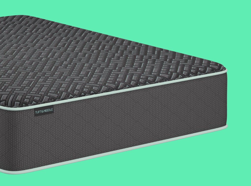 A close-up of a Tuft and Needle Mint Luxe mattress corner showing a dark textured top surface and a dark gray side panel with a small visible TUFT & needle label, set against a solid mint green background.