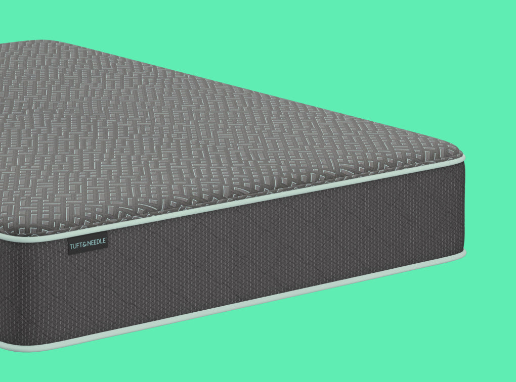 A close-up of a Tuft and Needle Original II mattress corner showing a textured gray top surface and dark charcoal side panel with a small visible TUFT & needle brand label, set against a solid mint green background.