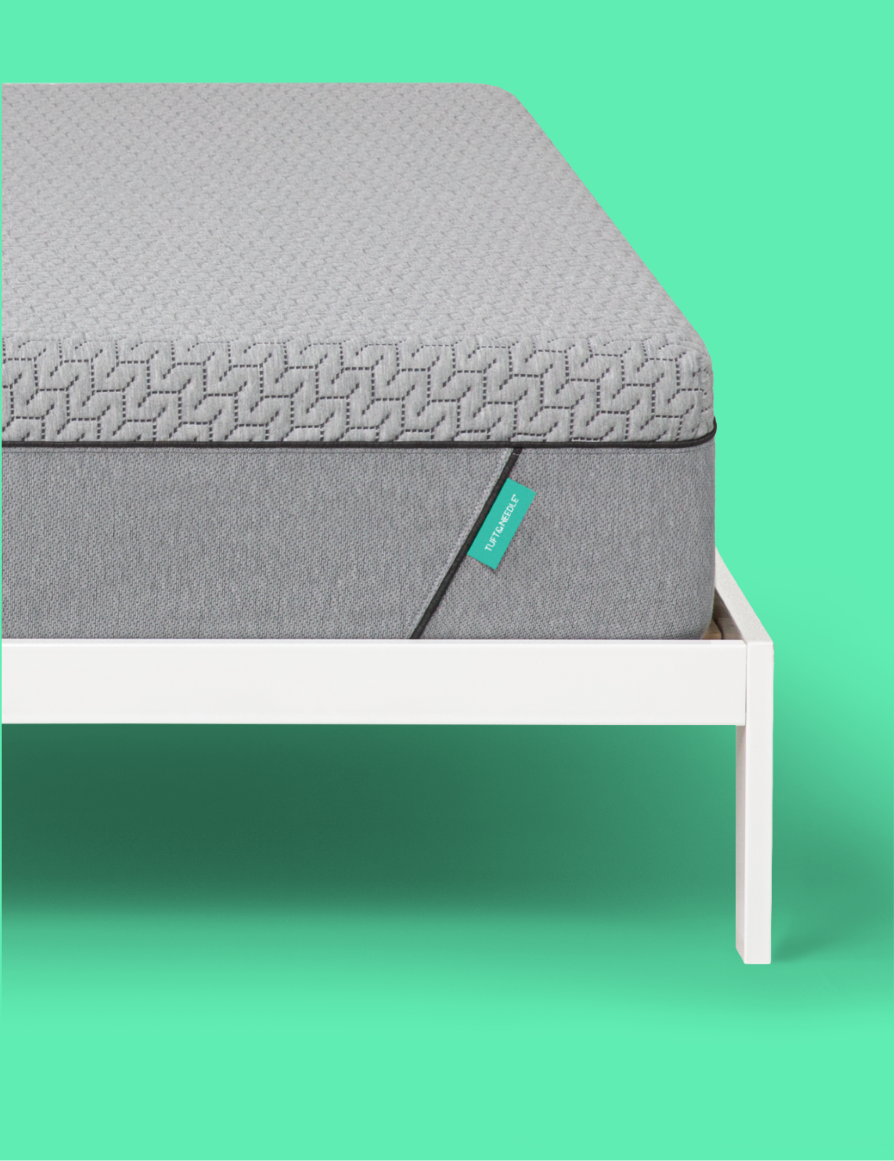 A close-up of a gray Tuft and Needle mattress on a white bed frame against a solid mint green background, with a small teal tag on the side of the mattress.