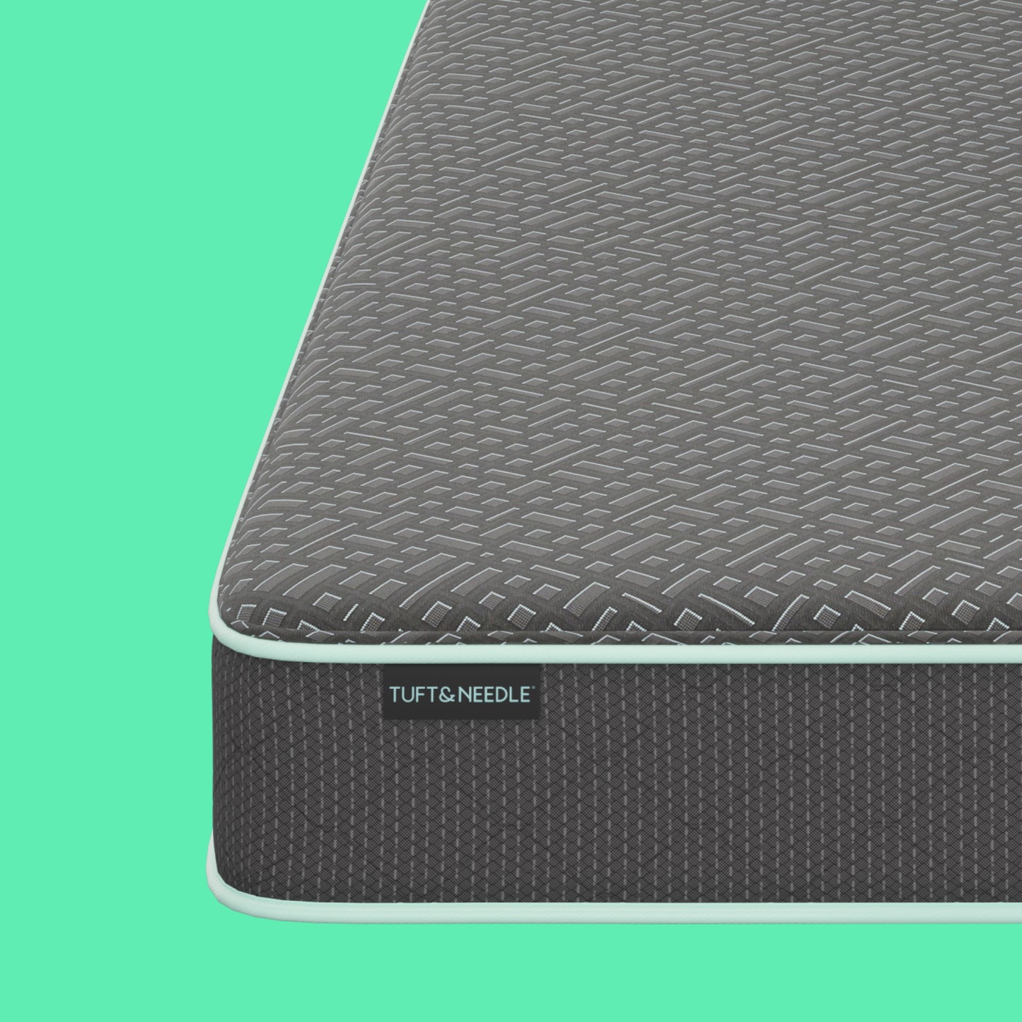 Close-up of a Tuft and Needle mattress corner showing a textured dark gray top surface and side panel with a visible TUFT & NEEDLE label, set against a solid mint green background.