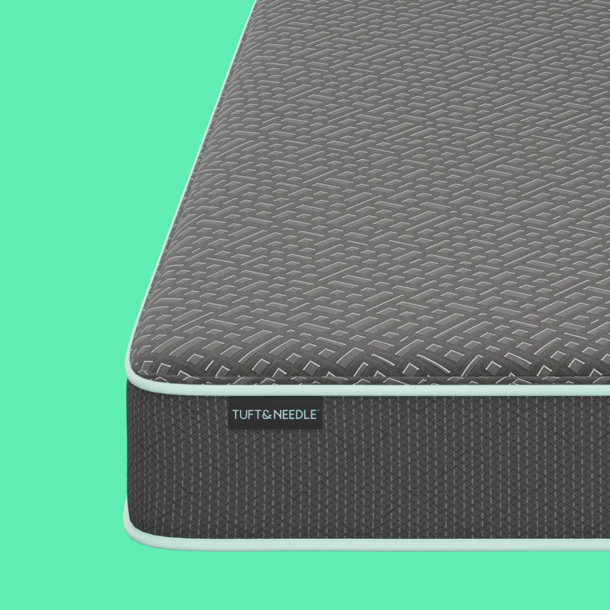 Close-up of a Tuft and Needle mattress corner showing a textured dark gray top surface and side panel with a visible TUFT & NEEDLE label, set against a solid mint green background.