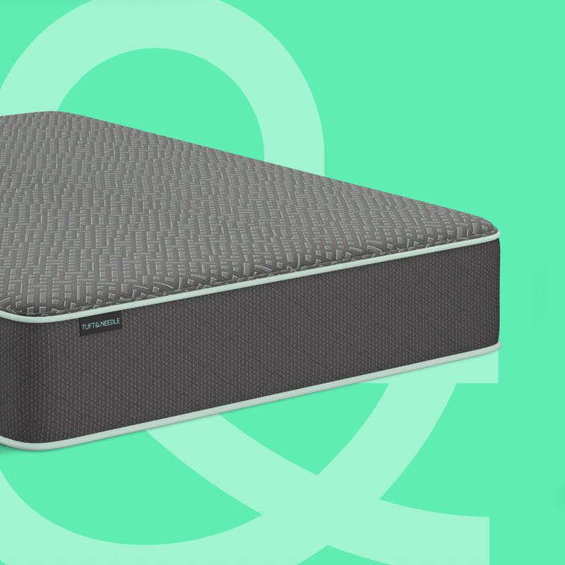 A dark gray Tuft & Needle 13 inch Mint mattress II shown at an angle against a mint green background with abstract light green shapes. The brand name Tuft & Needle is visible on a small label on the side of the mattress.