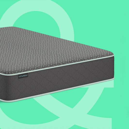 A dark gray Tuft and Needle mattress shown at an angle against a mint green background with abstract light green shapes. The brand name 'TUSKMEAT' is visible on a small black label on the side of the mattress.