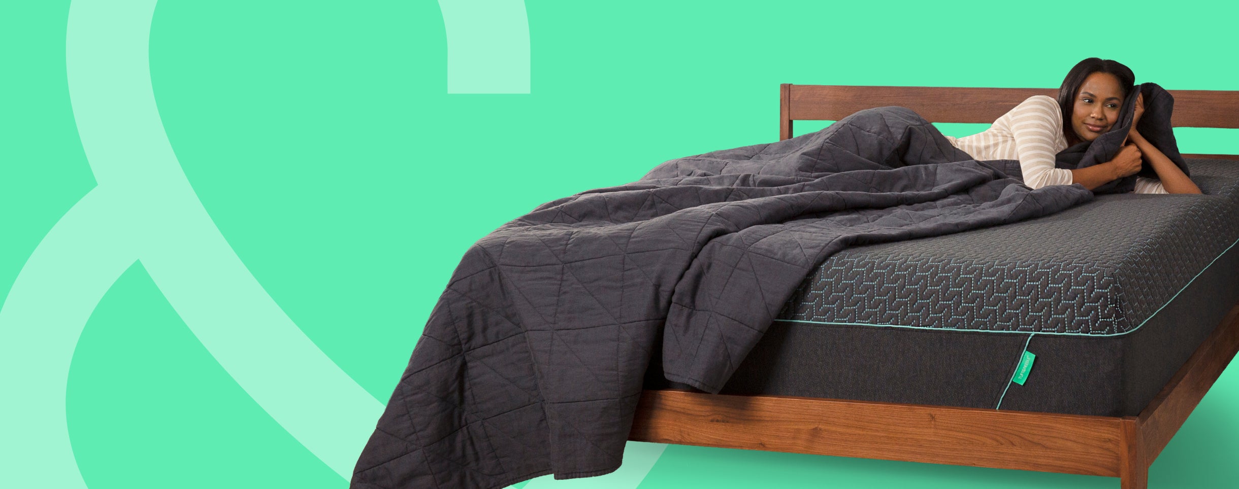 A woman lies on a Tuft & Needle mattress under a dark gray quilted comforter on a wooden bed frame, set against a bright mint green background with abstract white line art.