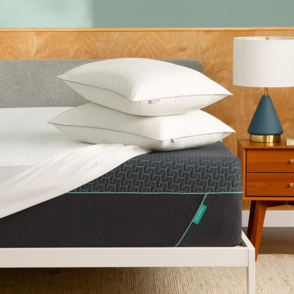 A dark gray Tuft and Needle mattress with a textured geometric pattern and a small teal tag, styled on a white bed frame with two stacked white pillows. A wooden nightstand with a blue and gold lamp is visible on the right, set against a wood-paneled wall and a teal accent wall.