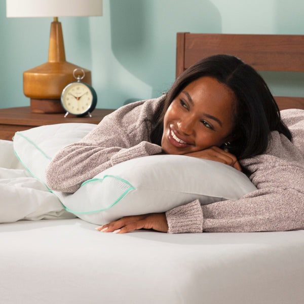 A smiling woman lies on a bed with white bedding, resting her head on a white Tuft and Needle Zip Comfort pillow with mint green piping. She wears a heathered taupe knit sweater.